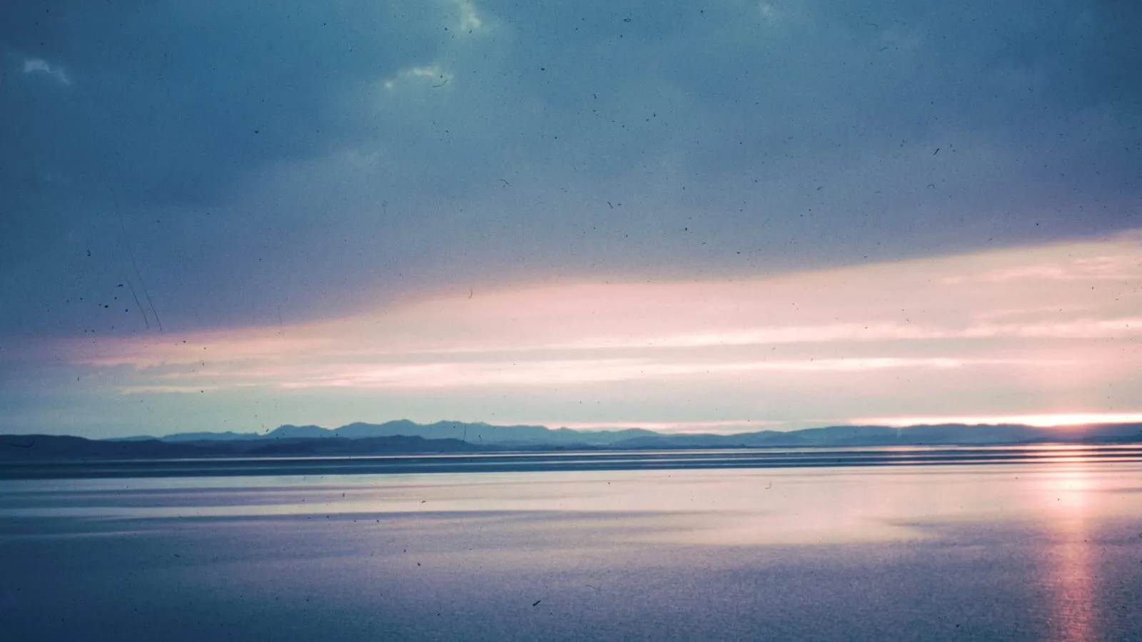 A cloudy sky shifting in tone above a calm lake at dawn, suggesting changing moods settling into stillness.