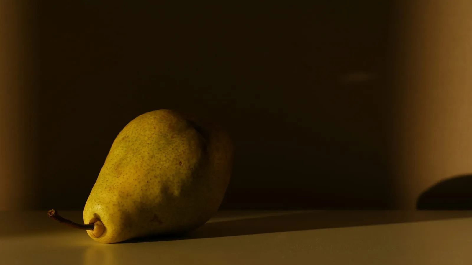 A single ripe pear resting on a plain table in soft natural light, a calm and simple still life.