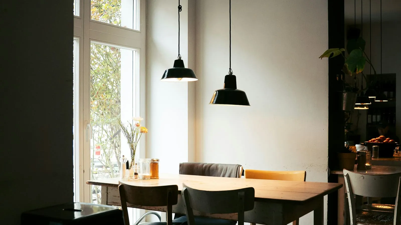 An empty cafe interior with tables and chairs beside a large window in soft daylight, suggesting a quiet social space.