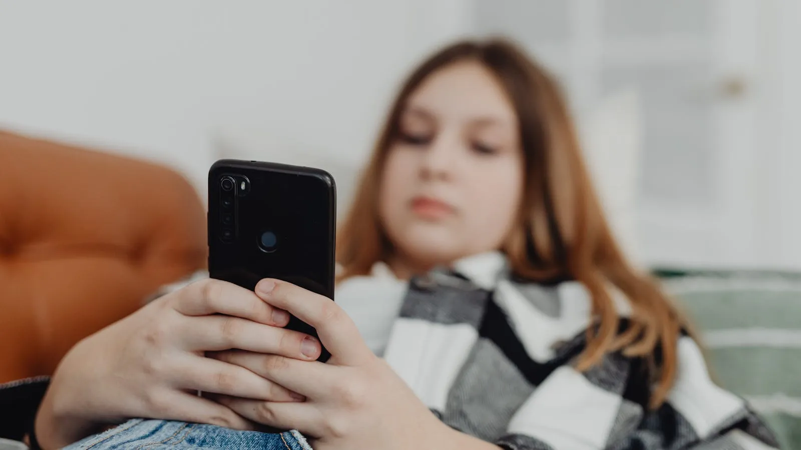 Teenager in a hoodie scrolling on a phone in a dim room, expression unreadable.