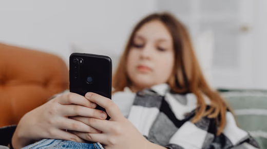 Teenager in a hoodie scrolling on a phone in a dim room, expression unreadable.