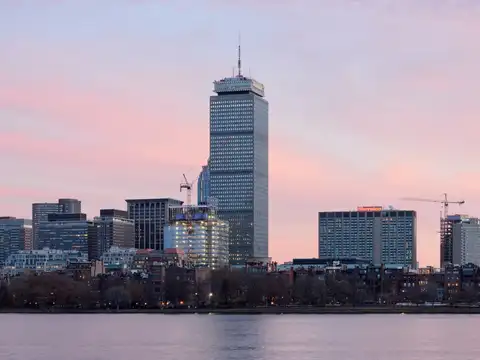 The Boston skyline at dusk seen across the Charles River, with the Prudential Tower rising above Back Bay under a pink sky.