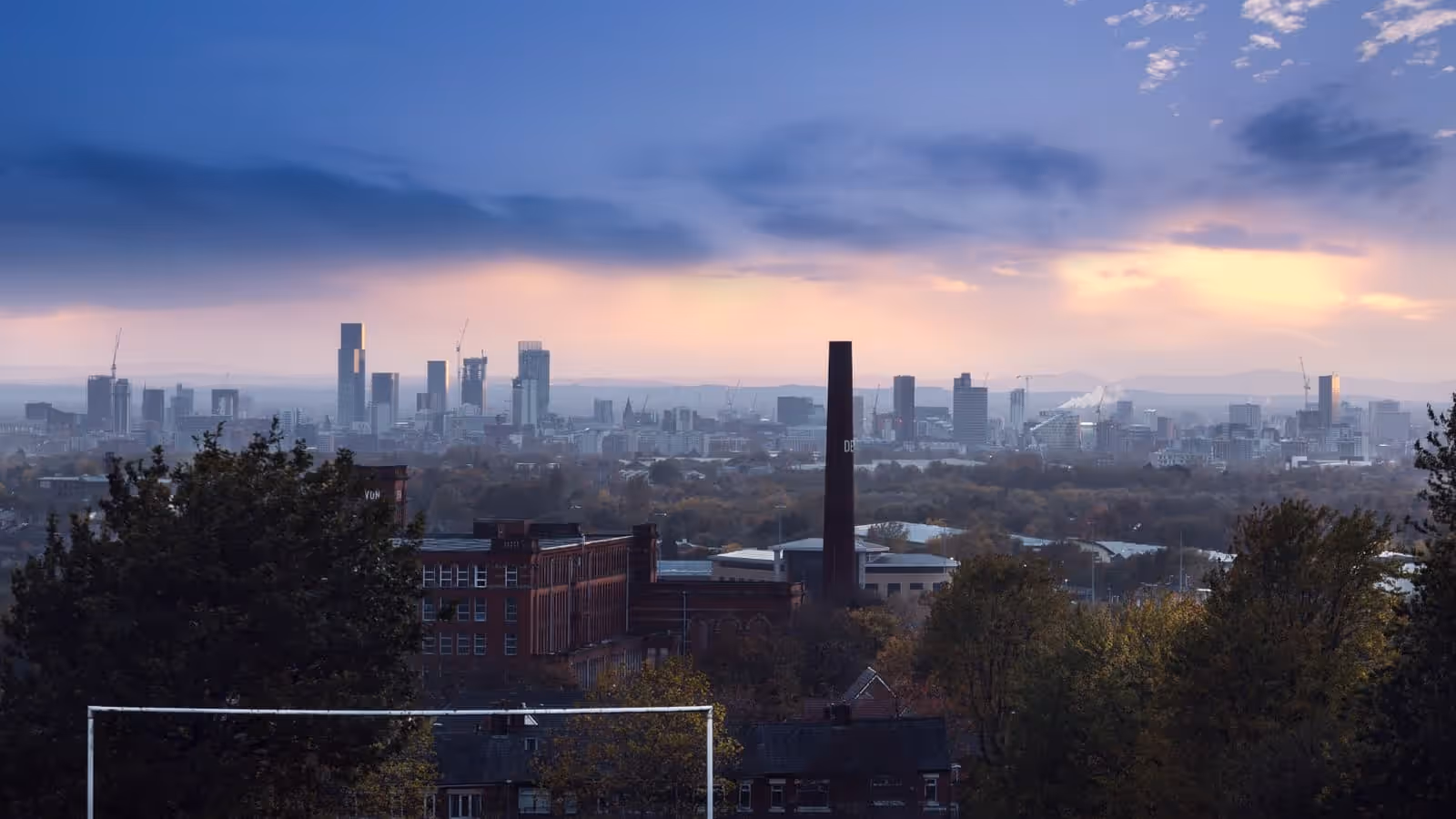 Die Skyline von Manchester bei Dämmerung, gesehen von einem erhöhten Punkt im Osten, mit den Türmen des Stadtzentrums am Horizont und einer roten Backsteinmühle sowie einem Schornstein im Vordergrund.