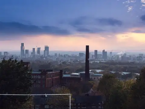 The Manchester skyline at dusk seen from high ground to the east, with the city centre towers on the horizon and a red-brick mill and chimney in the foreground.