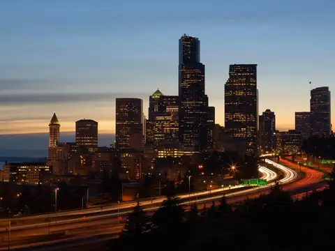 The downtown Seattle skyline at dusk, with the Columbia Center and the pyramid-topped Smith Tower above Interstate 5, light trails from traffic below and Elliott Bay beyond.