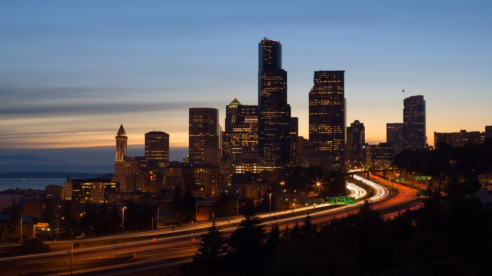 Die Skyline von Seattle im Stadtzentrum bei Dämmerung, mit dem Columbia Center und dem pyramidenförmigen Smith Tower über dem Interstate 5, Lichtspuren vom Verkehr darunter und der Elliott Bay im Hintergrund.